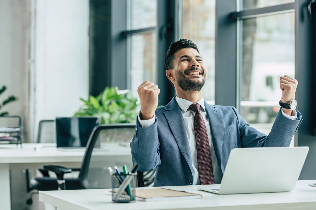 happy businessman looking up and showing winner gesture while sitting at workplace